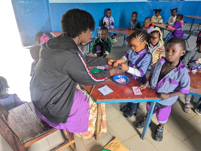 A classroom of young students sitting at small tables, with a teacher facing off camera speaking to one student. 