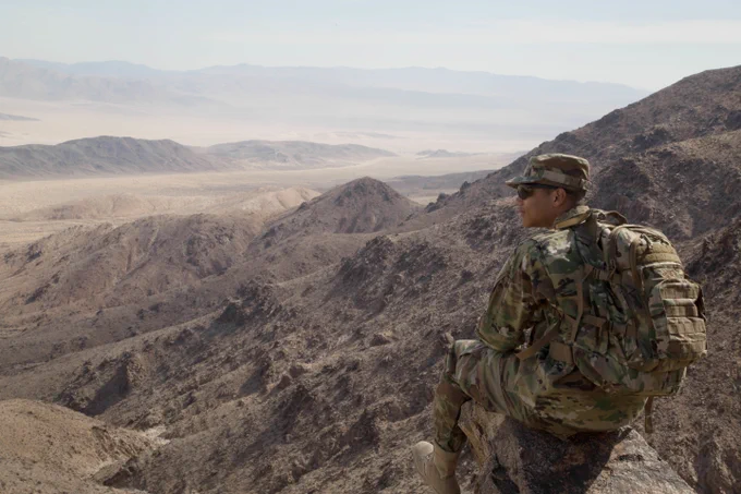 A Soldier in camouflage wearing a backpack and sunglasses sites on a rock in the mountains overlooking the desert floor. (Photo Credit: Staff Sgt. Douglas Lovely, 356th Broadcast Operations Detachment, U.S. Army)