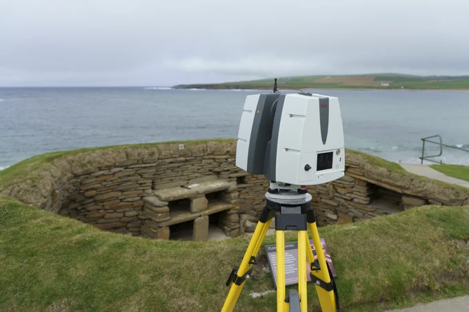 A laser scanner on a tripod looking at a low, historic stone structure at Skara Brae