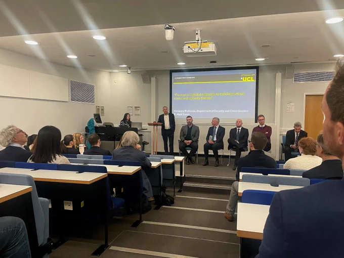 A group of attendees and speakers at the Five Eyes event in UCL Engineering's Lecture Theatre. A presentation slide is displayed at the front, where several individuals are seated on chairs facing the audience. The room features standard lecture seating, presentation equipment, and white walls
