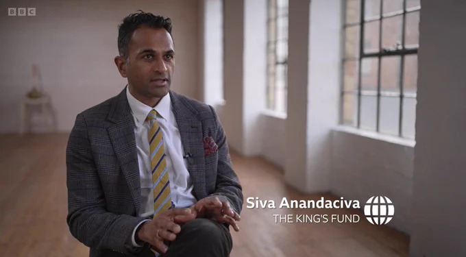 Image of Siva Anandaciva sat in a large open plan room, with tall windows to the right-hand side of the image. 