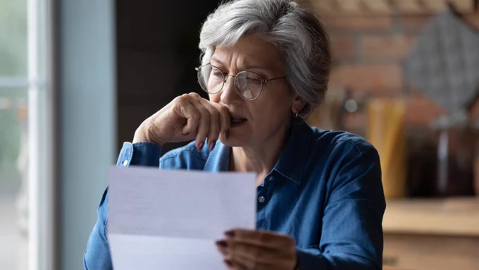 A nervous looking woman reading a letter in her kitchen.
