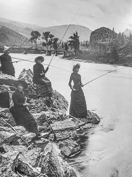 A black and white image of women wearing long dark dresses and hats stand fishing along the edge of a river in the mountains.