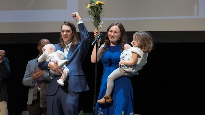 Two people on a stage, each holding a child, celebrating a victory with one of them raising a bouquet of flowers, as the audience claps.