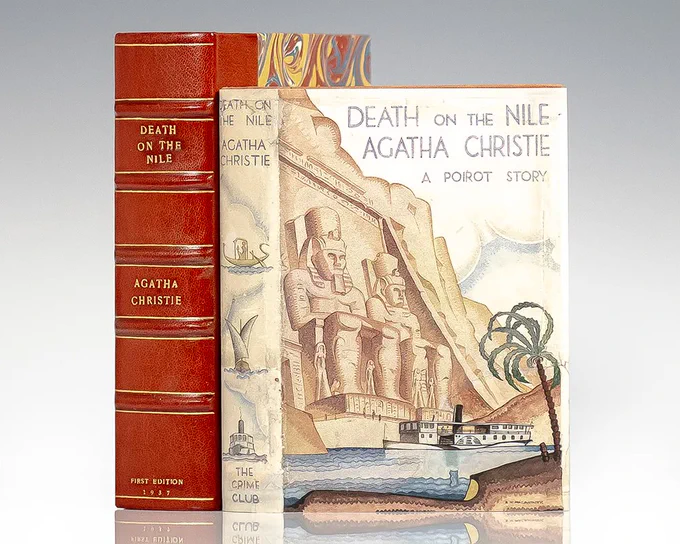 A vintage first edition of 'Death on the Nile' by Agatha Christie from 1937, displayed showing both the red leather-bound spine with gold lettering and the dust jacket. The cover art depicts Egyptian statues at Abu Simbel with a steamboat cruising on the Nile River below, palm trees along the shore, and Egyptian-inspired illustrations in a watercolor style.