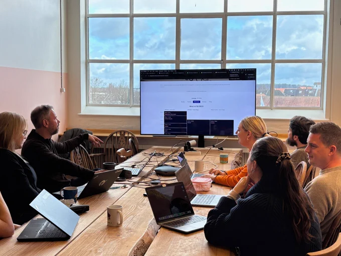 Six professionals gathered around a wooden conference table in a brightly lit room, focusing on a large monitor displaying a web interface. The room features a large window with a view of a cloudy sky and distant buildings. The group is engaged in a discussion led by a man pointing at the screen, indicating a collaborative and productive meeting atmosphere.