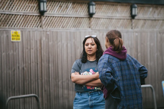 Two figures stand, outside, on location, in front of a fence, in conversation. One with her  back to the camera, one facing. The person facing the camera is Fozia O'Dowd - a brown woman with long black hair, arms folded, wearing a t-shirt and jeans with sunglasses on her head 