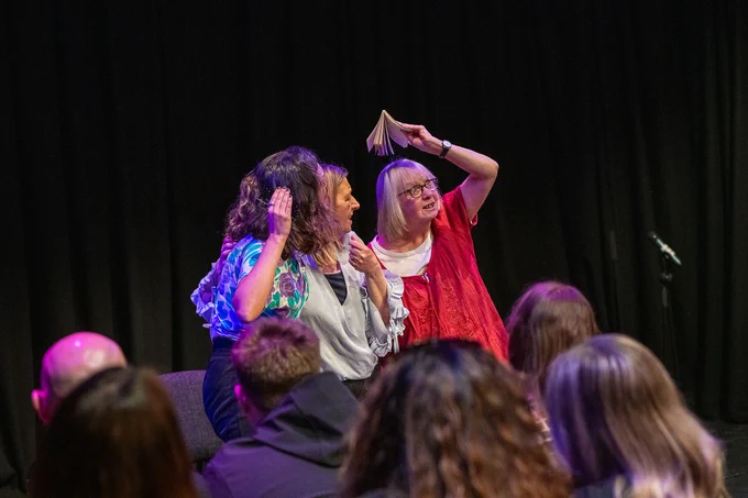 Three people on stage performing a scene, with others watching them, one of the women on stage is holding a book above her head as if a hat to shelter herself 