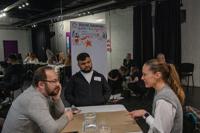 Three people sat at a table talking to each other, with Creative Placemaking banner behind them. There are others sitting at different tables around the room, participating in the workshop 