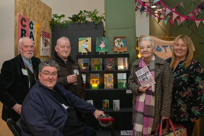 Group of people in front of a display of artwork, one of them is holding a Craft Cafe newsletter, theyre looking into the camera. 