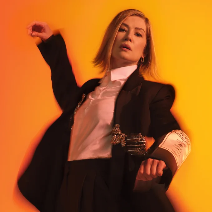 Person in a stylish black suit and white shirt, posing dynamically against an orange background.