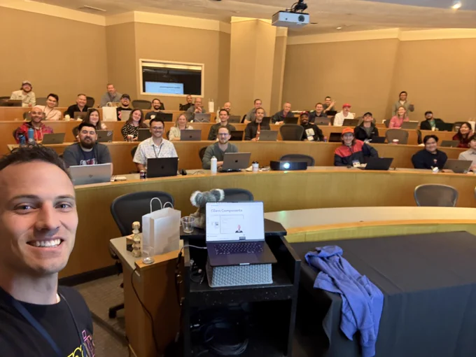 A selfie taken by a smiling instructor at the front of a tiered classroom filled with attendees, all seated with laptops and looking toward the camera. A laptop on a podium displays a slide titled “Client Components.” Nearby are EpicWeb-branded swag items including a gift bag, wooden stacking blocks, and a stuffed koala. The atmosphere is cheerful and focused, suggesting an engaging tech workshop or conference session.