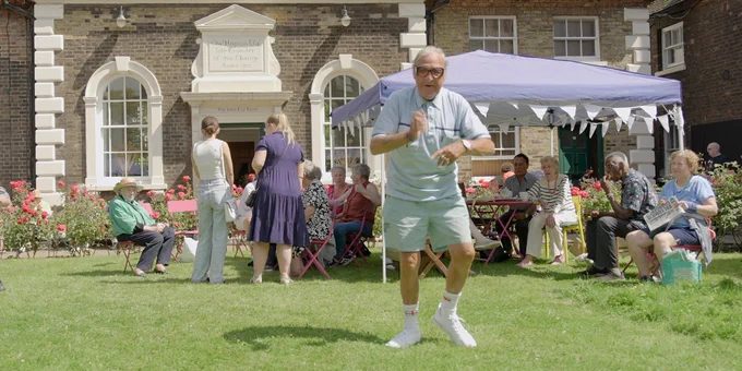 An individual is dancing enthusiastically in the foreground at an event outside, while several others sit and talk in the garden.