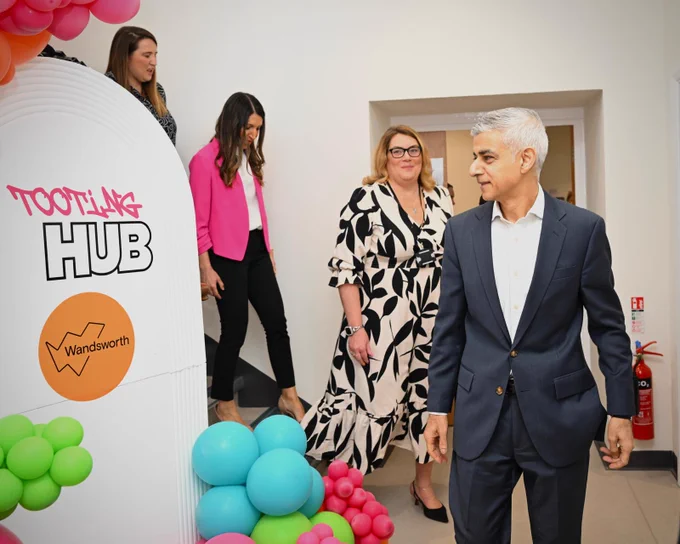 Mayor of London is smiling as he walks through Tooting Hub, followed by three people. A white sign with colourful balloons marks the venue entrance - the sign says “Tooting Hub Wandsworth”.