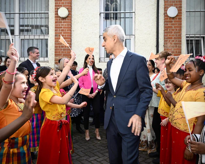Mayor of London outside Tooting Hub - greeted by young Londoners. Everyone is smiling and the young people are waving mini flags. 