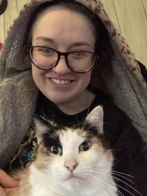 Photo of a white Hispanic nonbinary person with glasses and smiling, wearing a leopard print hooded robe with a beautiful white calico cat in front of them.