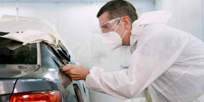 A person in protective clothing and goggles inspecting the damage on a car in a collision repair shop.