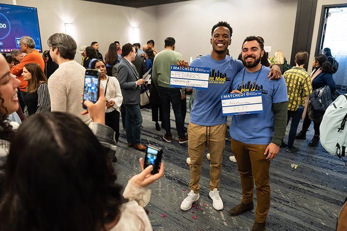 Two smiling medical students stand arm-in-arm in a crowded room celebrating Match Day. They wear matching blue Pitt Med Class of 2024 shirts and hold signs showing their residency matches—one in Pediatrics at Nationwide Children’s and the other in Anesthesiology at Pitt. Around them, friends and family take photos with their phones, capturing the joyful moment. The room is filled with people mingling and celebrating, with confetti scattered on the carpeted floor.