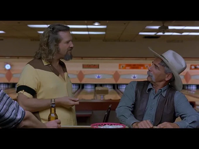Seated at the bowling alley bar, the Stranger looks up at the standing Dude as the two men converse.