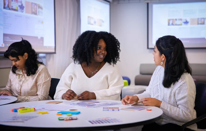 3 women sitting at desk with paper and highlighter pens. 2 are in conversation