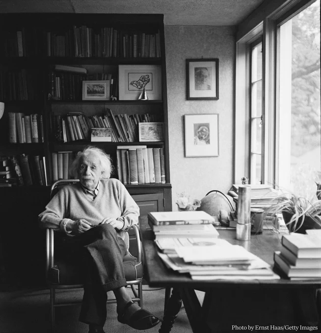 Albert Einstein sitting in a chair beside a desk with papers, with bookshelves behind. 