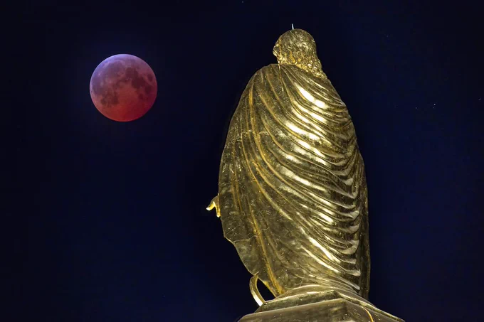 View of the back of the golden statue of the virgin Mary atop the Main Building at the University of Notre Dame under an ink-black night sky, with a total lunar eclipse featuring a reddish moon visible in the background.
