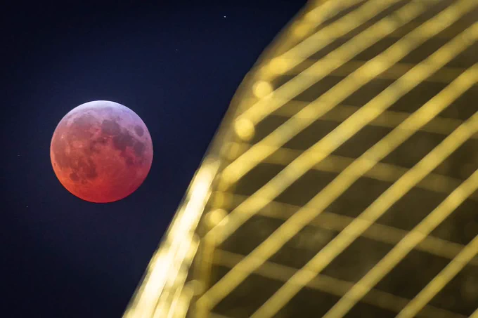 A lunar eclipse with a red moon next to the curved, illuminated facade of the Golden Dome at the University of Notre Dame.
