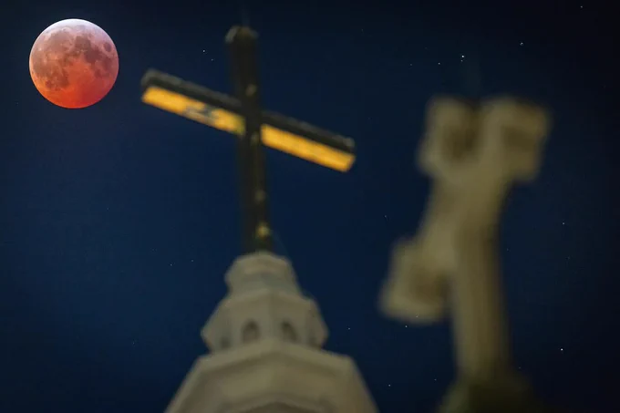 A lunar eclipse beside a church steeple topped with a cross, under a starry sky.