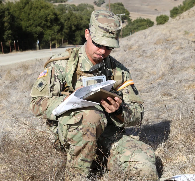 A photo of a kneeling Soldier holding a map and a protractor. (Photo Credit: Spc. Elizabeth Buck, 69th Public Affairs Detachment, U.S. Army National Guard; modified by the Army Geospatial Center)