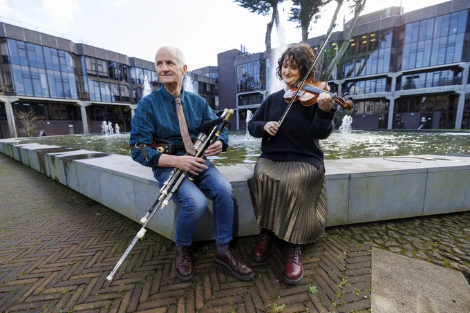 Photograph of a man on the left playing uilleann pipes next to a woman in the right playing the fiddle. They are sat on the edge of a fountain with large glass-fronted buildings in the background
