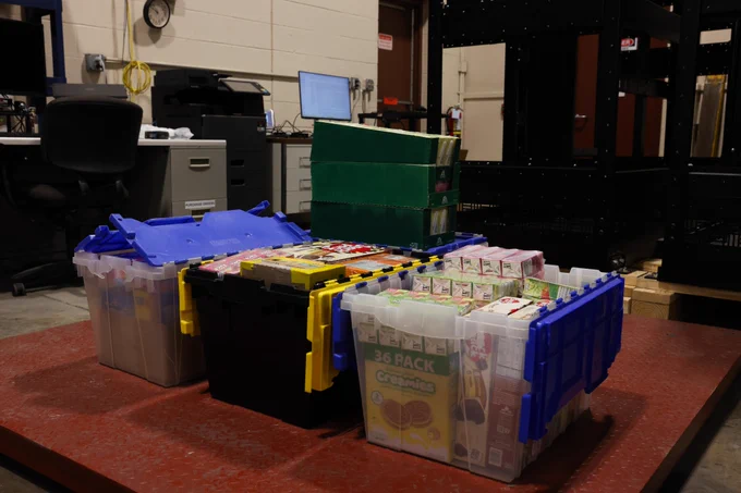 This image shows food items inside of four plastic containers and three packages of juice boxes stacked on top of one of the plastic containers. These containers and boxes are sitting on top of a scale. 