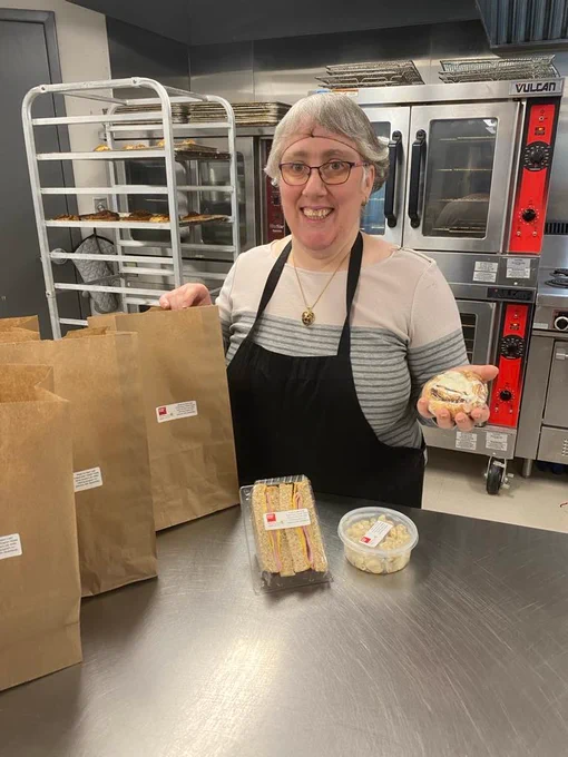 A woman wearing an apron stands in a commercial kitchen, smiling at the camera. She holds a cinnamon bun in one hand and is surrounded by brown paper bags filled with baked goods. In front of her, there are two containers: one with a ham and cheese sandwich and one with pasta salad.