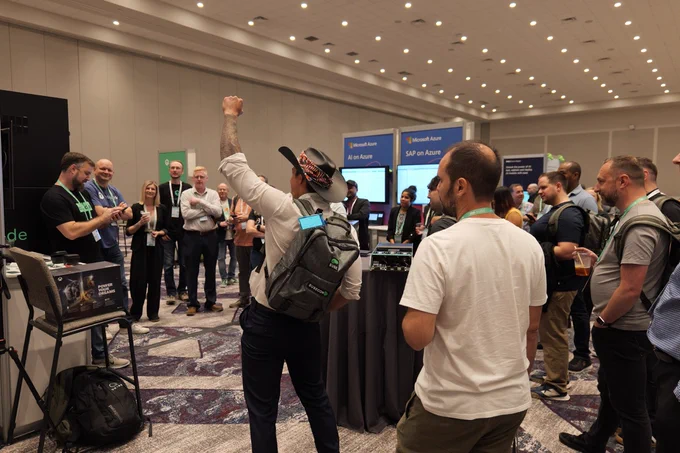 A conference booth scene where a person wearing a backpack and a cap raises his fist in celebration, surrounded by attendees who are clapping and smiling. The booth in the background displays the “Oxide” logo, and additional signage referencing Azure services is visible in the large, well-lit convention hall.