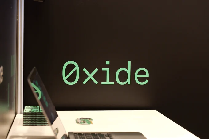 A close-up of the word “Oxide” in green text on a black backdrop. In front of the backdrop, a white table holds a partially visible laptop and a small circuit board.