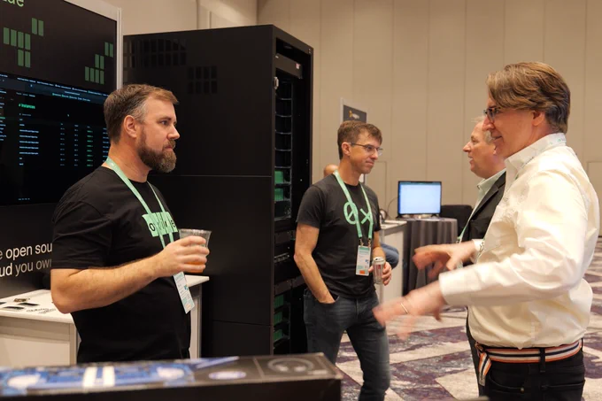 A group of people conversing at a technology conference booth. One man in a black T-shirt stands beside a tall server rack branded with “Oxide,” holding a drink while talking with two other attendees.