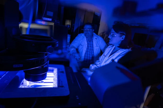 Two scientists examining a sample under a microscope in a dark laboratory illuminated by blue light.