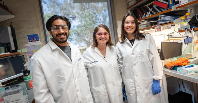 Three researchers wearing lab coats stand in a laboratory, with various scientific equipment in the background. They each have a badge with the logo of UC San Diego visible on their coats.