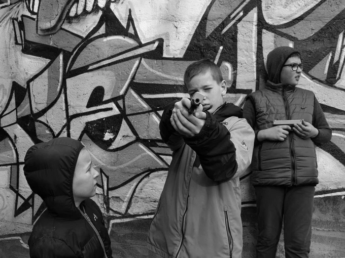 A boy points a replica gun straight at the camera. His expression is a mix of playfulness and intensity, blurring the line between childhood and harsh reality.