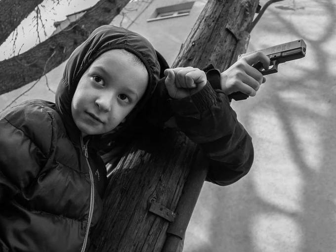 A young boy in a hoodie holds a replica assault rifle. His face is serious, and the background of crumbling walls reflects an environment filled with uncertainty.