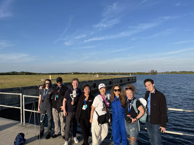 A group of eight NASA Social participants smile for a group photo at Kennedy Space Center in daytime with blue skies above. They are standing at a wharf with a metal fence and water behind them and a launch pad in the distance.