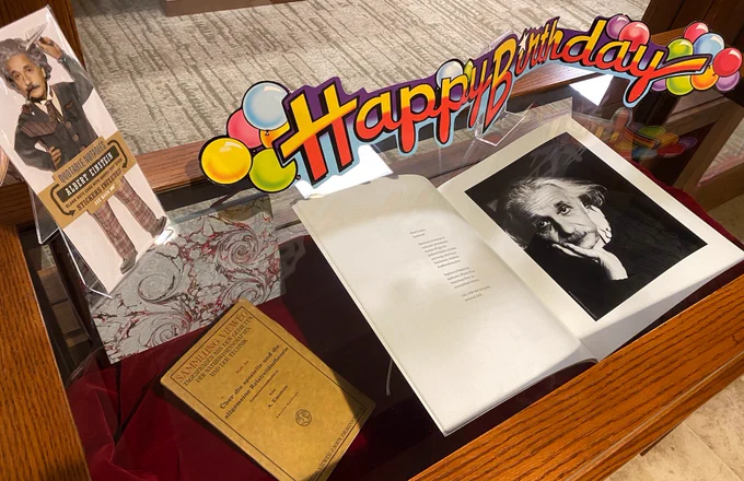 Photo of display case with several items, including large book with Einstein photograph, smaller work on relativity, UnEmployed Philosopher’s Guild Einstein figure, and Happy Birthday sign.