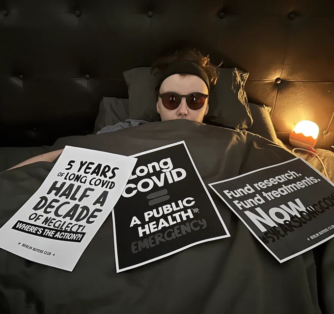 Photo of myself, a white woman, in bed with sunglasses and a small lamp. Three signs are laid on the bed which read “5 years of long Covid, half a decade of neglect. Where’s the action?”  “Long covid = a public health emergency” and “fund research. Fund treatments. NOW”