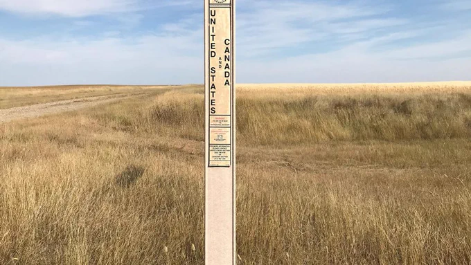The photo shows a grassy, open landscape with a clear blue sky dotted with a few wispy clouds. The terrain appears to be a mix of tall, dry grass and patches of dirt, suggesting a rural or prairie setting. A narrow dirt path or road stretches across the scene from left to right, dividing the landscape.
In the foreground, there is a vertical marker or signpost standing in the grass. The sign has text on it, with the most prominent words being "UNITED STATES AND CANADA" written vertically in large, bold letters. Below this, there is smaller text, likely providing additional information, but it's not fully legible in the image. 