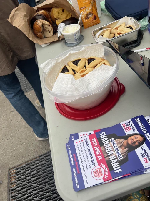 Photo of a table with campaign lit for Shahana Hanif next to a Tupperware full of hamantaschen.