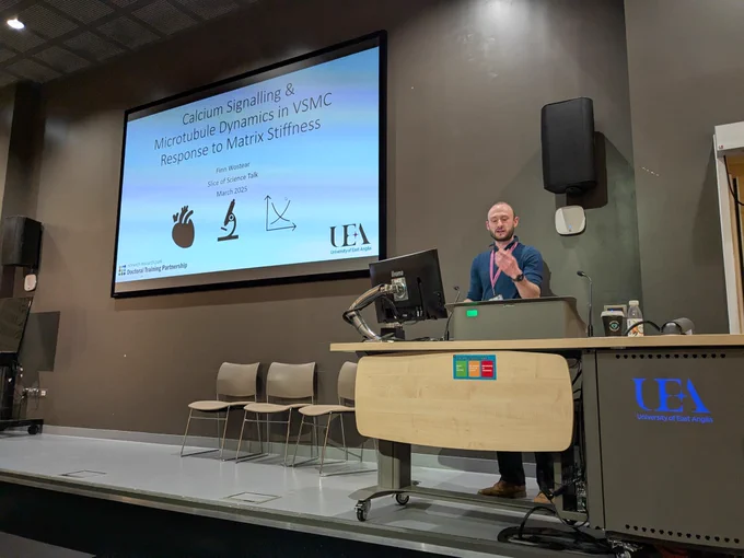 Speaker Finn Wostear stands behind a podium in a modern lecture hall, gesturing with one hand. He is wearing a dark blue sweater over a light-colored collared shirt, a pink lanyard, and brown shoes. A large screen behind him displays a PowerPoint slide with the title 'Calcium Signalling & Microtubule Dynamics in VSMC Response to Matrix Stiffness,' presented by Finn Wostear for a 'Slice of Science Talk' in March 2025. The slide includes the University of East Anglia (UEA) logo and three scientific icons: a heart, a microscope, and a graph. The podium has a built-in monitor and microphone, and a UEA logo is illuminated on the front.
