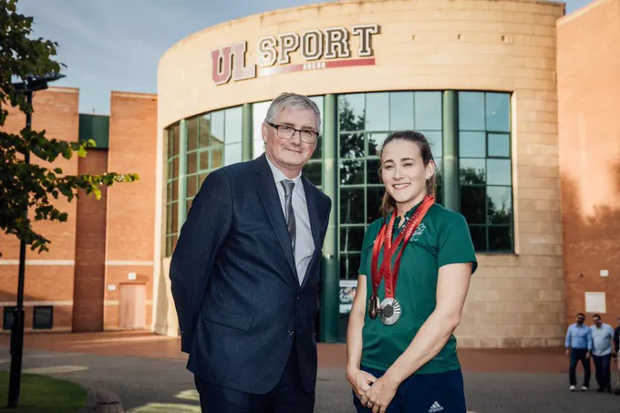 Photograph of a man in a blue suit on the left stood next to a woman in a dark green top with medals on red ribbons around her neck on the right. They are stood outside a large brick building with a "UL Sport" sign on it 
