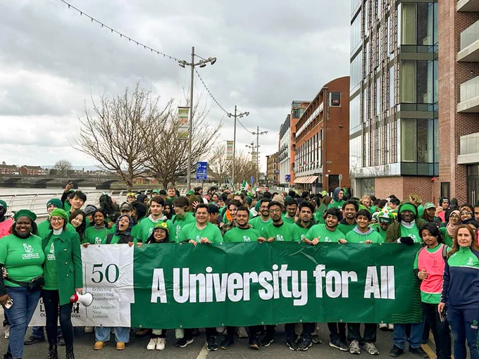 Photograph of a large group of University of Limerick students in green t-shirts gathered behind a banner which reads "A University for All". They are part of the 2025 Limerick St Patrick's Day parade