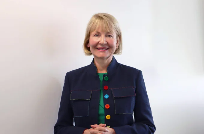 A professional portrait of a woman with short blonde hair, smiling at the camera. She is wearing a navy blue jacket with colorful buttons and stitching, over a green top. The background is plain white, and she stands with her hands gently clasped in front of her.







