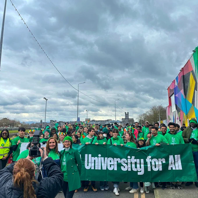 A large group of University of Limerick students gathered for the St Patrick's Day parade, all dressed in green t-shirts behind a dark green banner reading "A University for All"