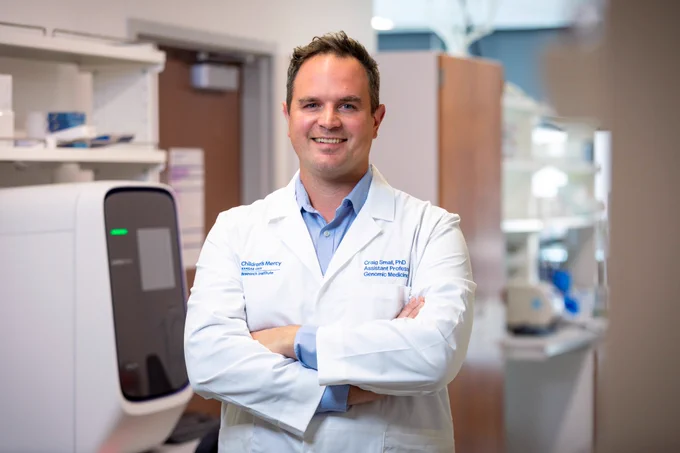 A photo of Dr. Craig Smail. He stands in the middle of a lab, wearing a white lab coat, with his arms crossed over his chest. He is smiling at the camera. You can see equipment behind him."